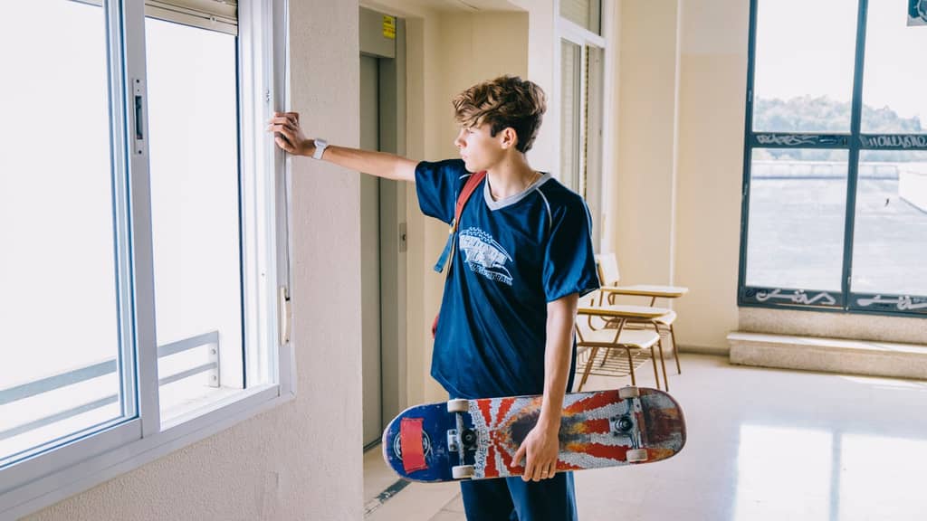 Boy holding a skateboard, looking out a window in a bright indoor setting, emphasizing the connection between indoor spaces and outdoor activities.