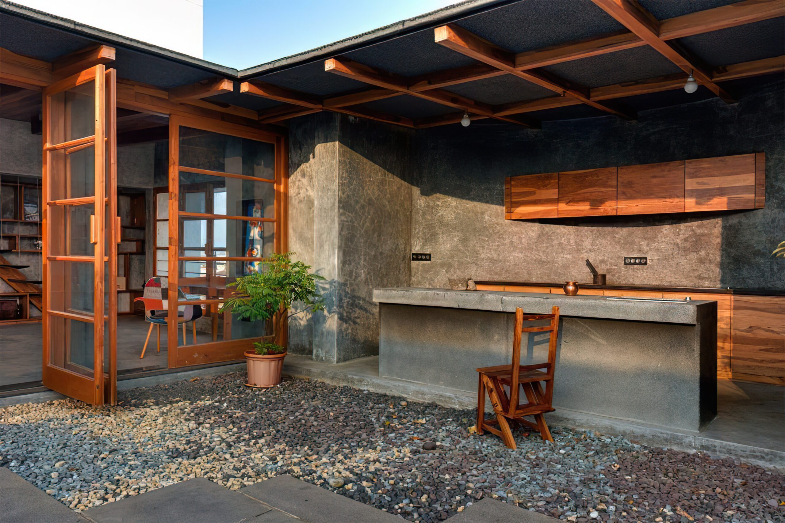 Custom outdoor kitchen with concrete countertop, wooden cabinetry, and folding wooden chair, surrounded by pebbled landscaping, showcasing modern design elements for outdoor living.