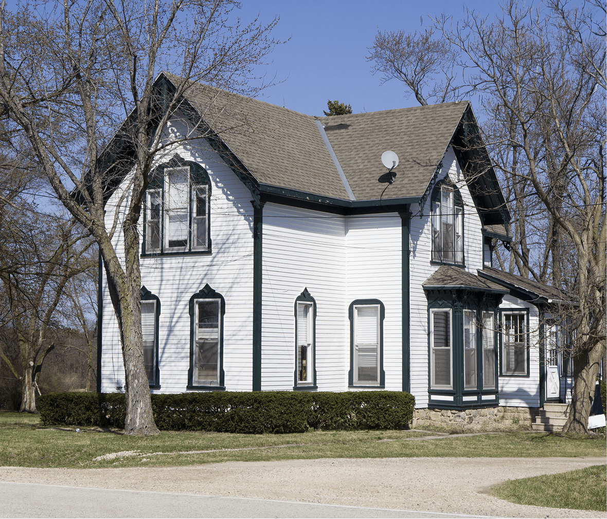 House with a white exterior and green trim, featuring multiple windows and a steep roof, surrounded by trees and manicured shrubs, reflecting a classic architectural style suitable for outdoor living spaces.