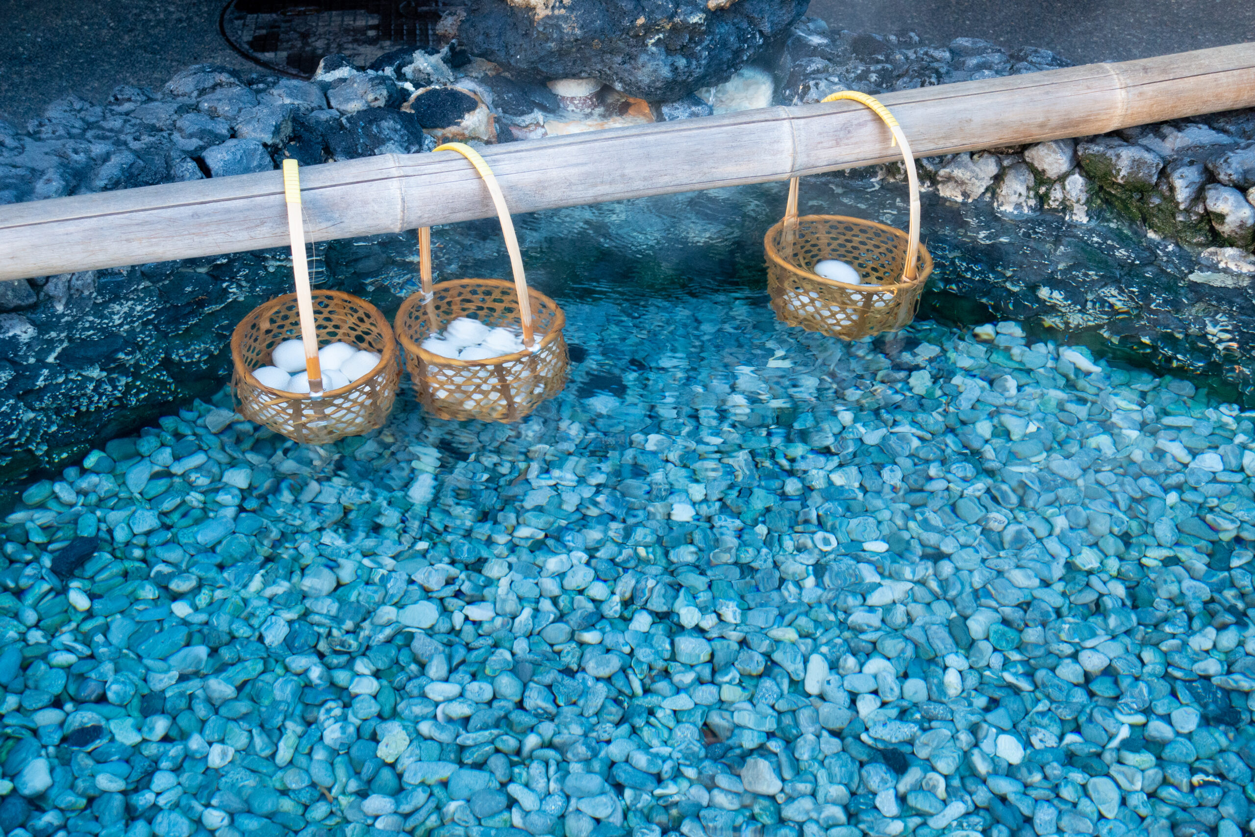 Baskets containing boiled eggs submerged in a clear hot spring, surrounded by smooth pebbles, showcasing a tranquil outdoor relaxation feature.