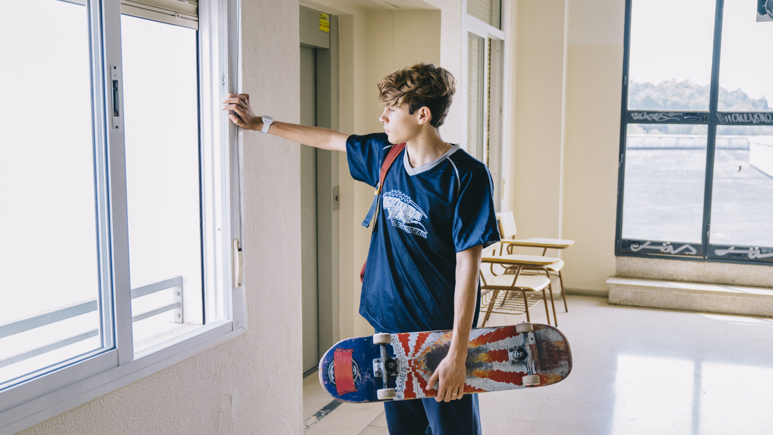 Boy holding a skateboard, looking out a window in a bright indoor setting, emphasizing the connection between indoor spaces and outdoor activities.