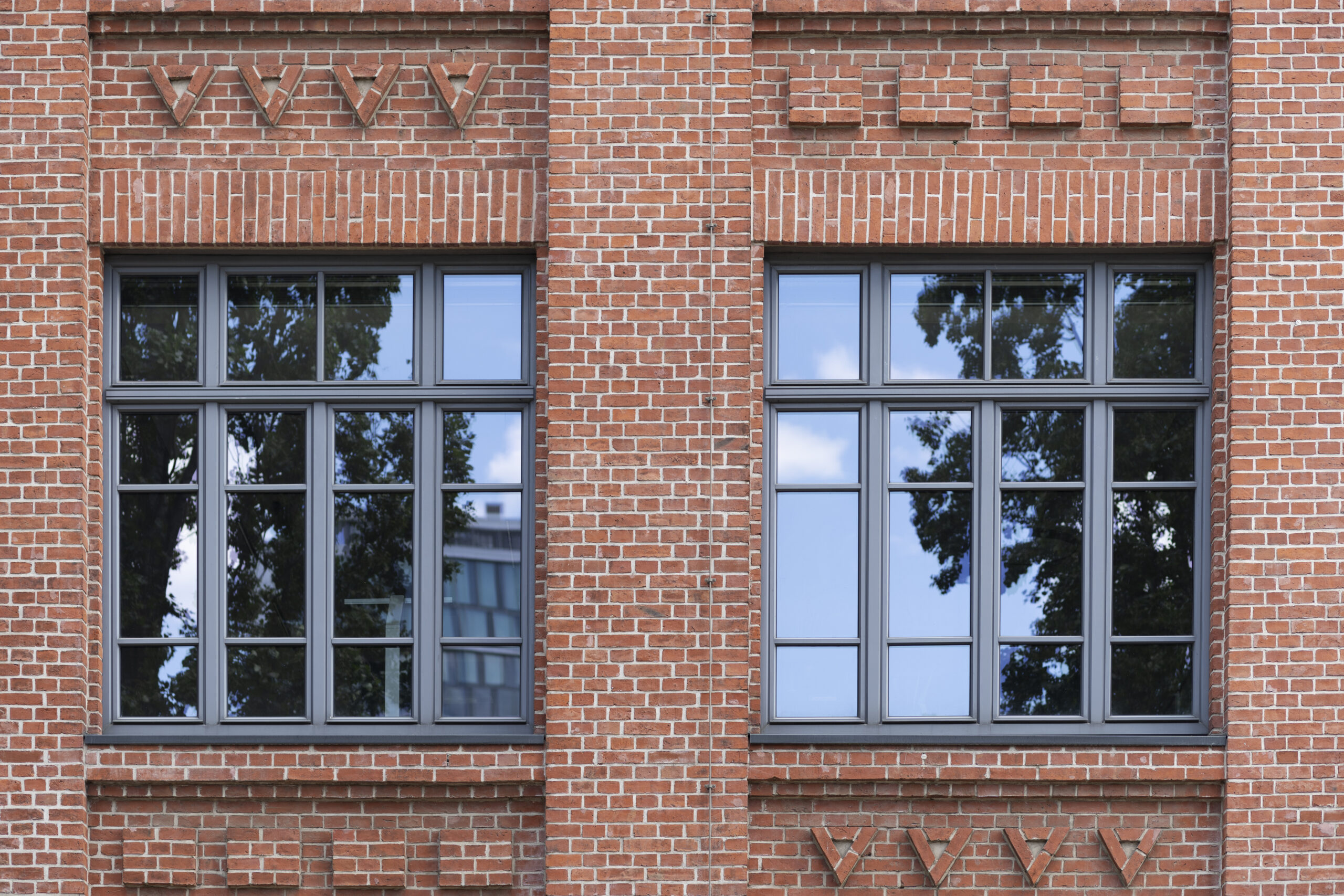 Pella 1800 Series Impact Windows installed on a brick wall, showcasing durability and energy efficiency, reflecting surrounding trees and sky.