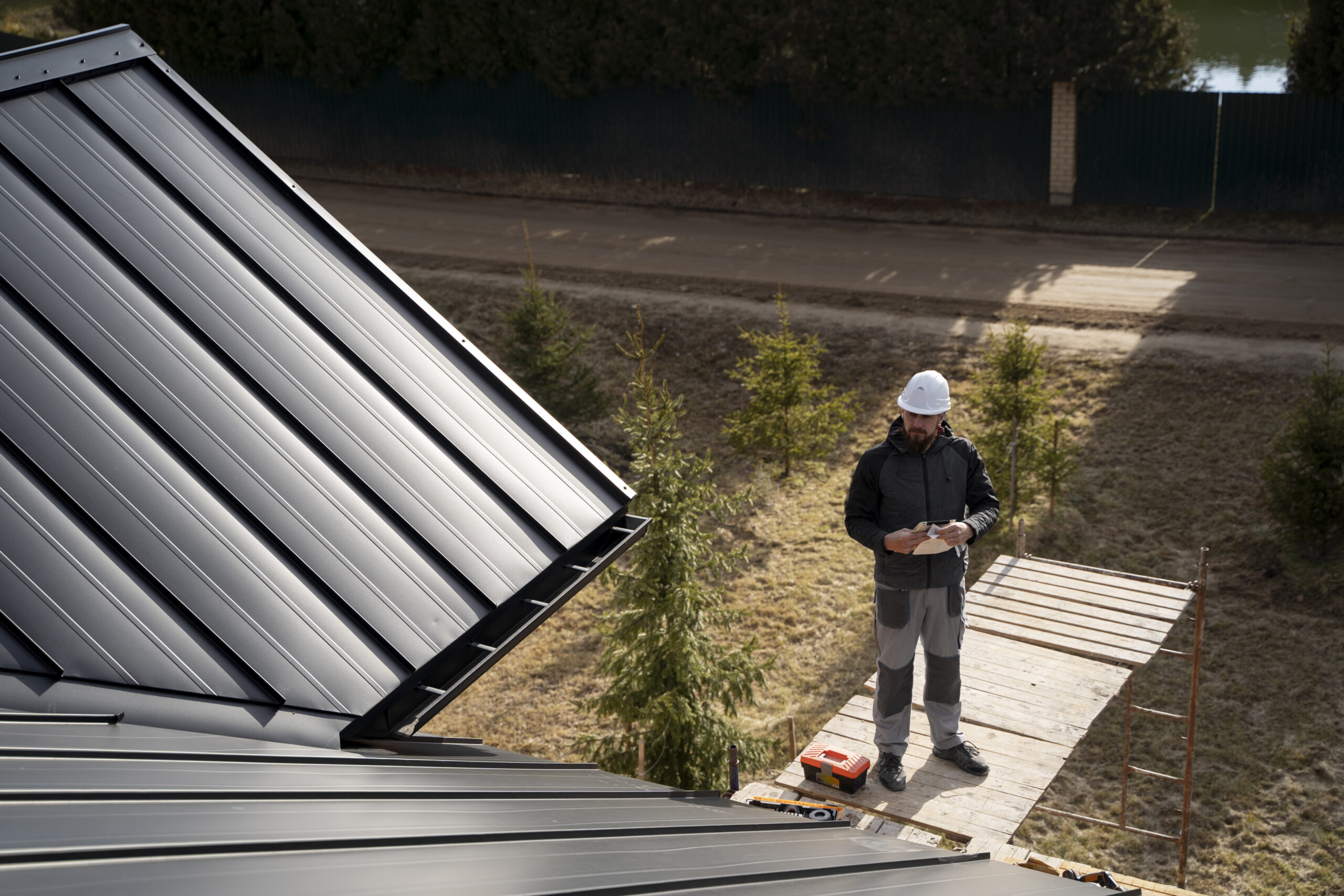 Man wearing a safety helmet standing on a scaffold, inspecting a metal roof installation, surrounded by trees and a roadway, emphasizing roofing expertise and safety in Oxford, MS.