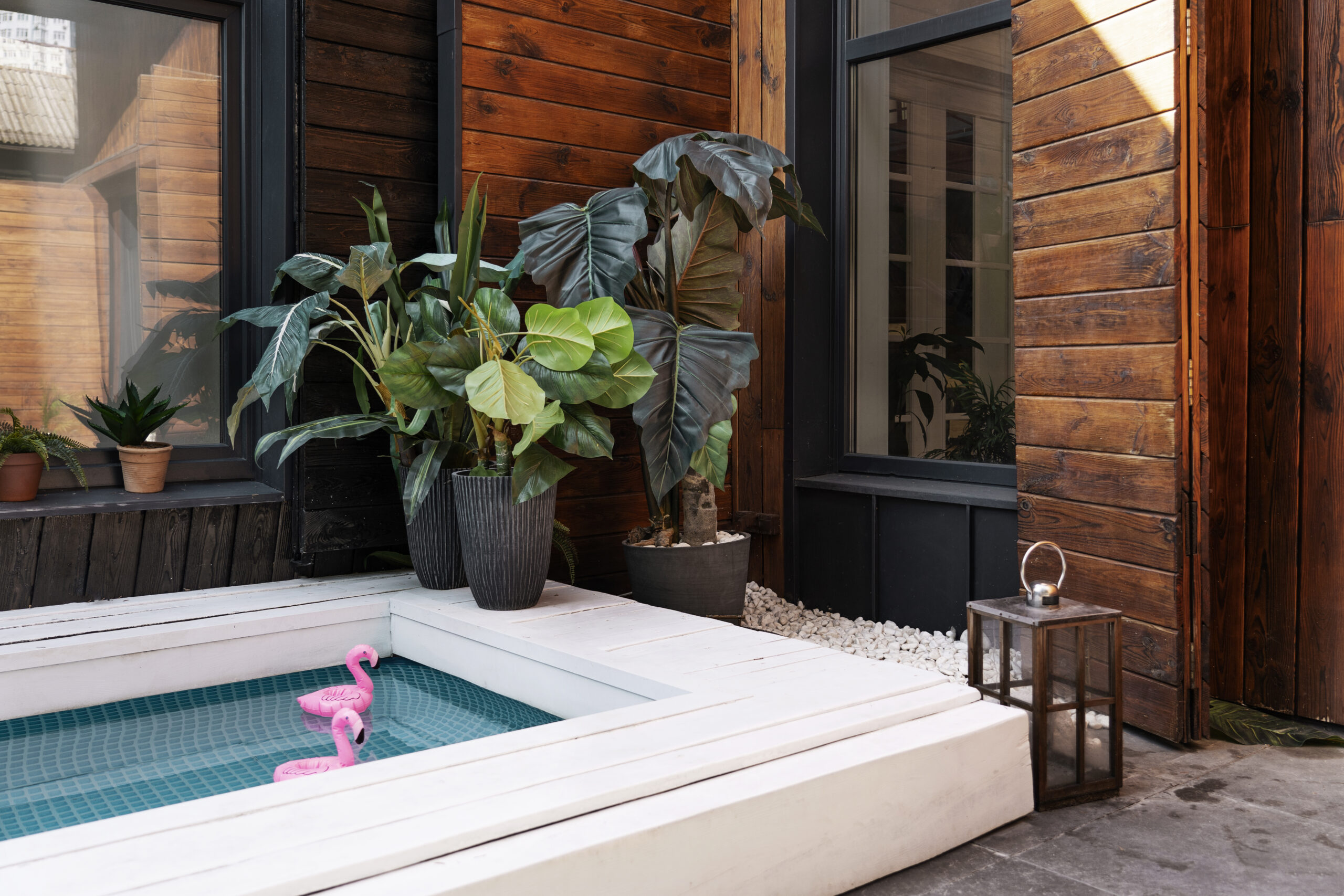 Outdoor spa area featuring a modern design with a small pool, lush green plants, and decorative elements like pink flamingo pool floats and a lantern, emphasizing relaxation and luxury in an outdoor setting.
