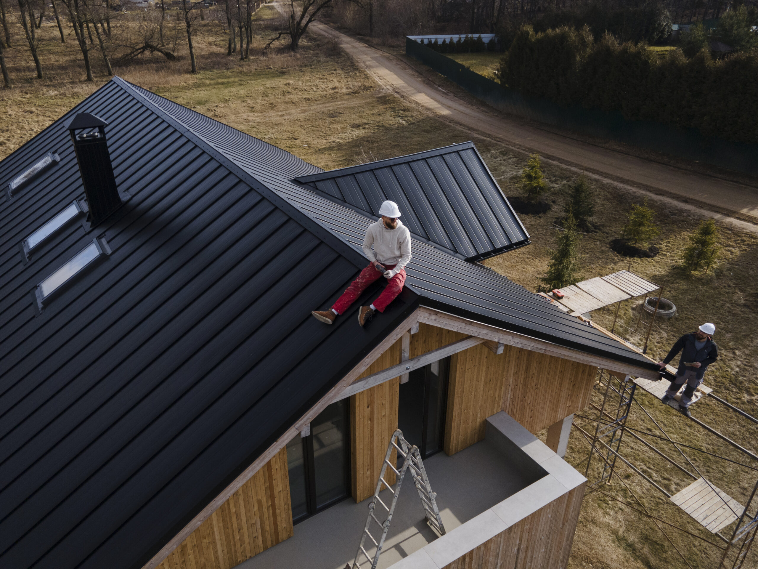 Man in helmet sitting on a black metal roof, overseeing installation, with another worker on scaffolding, showcasing residential roofing project by Tekton Exteriors in Oxford, MS.