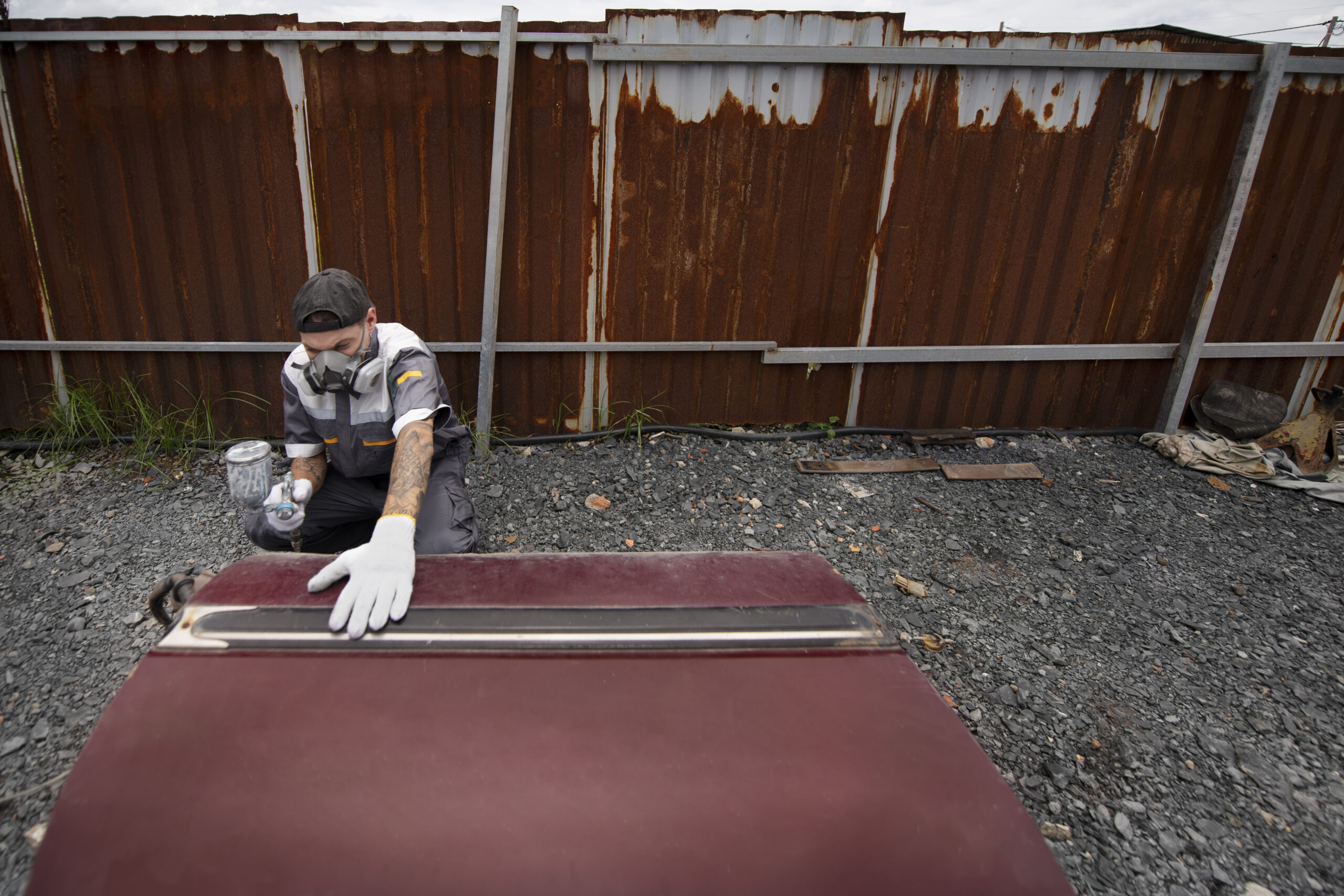 Man in protective gear applying a coating to a roof panel outdoors, surrounded by a rusted metal fence and gravel, emphasizing storm damage repair and restoration services.