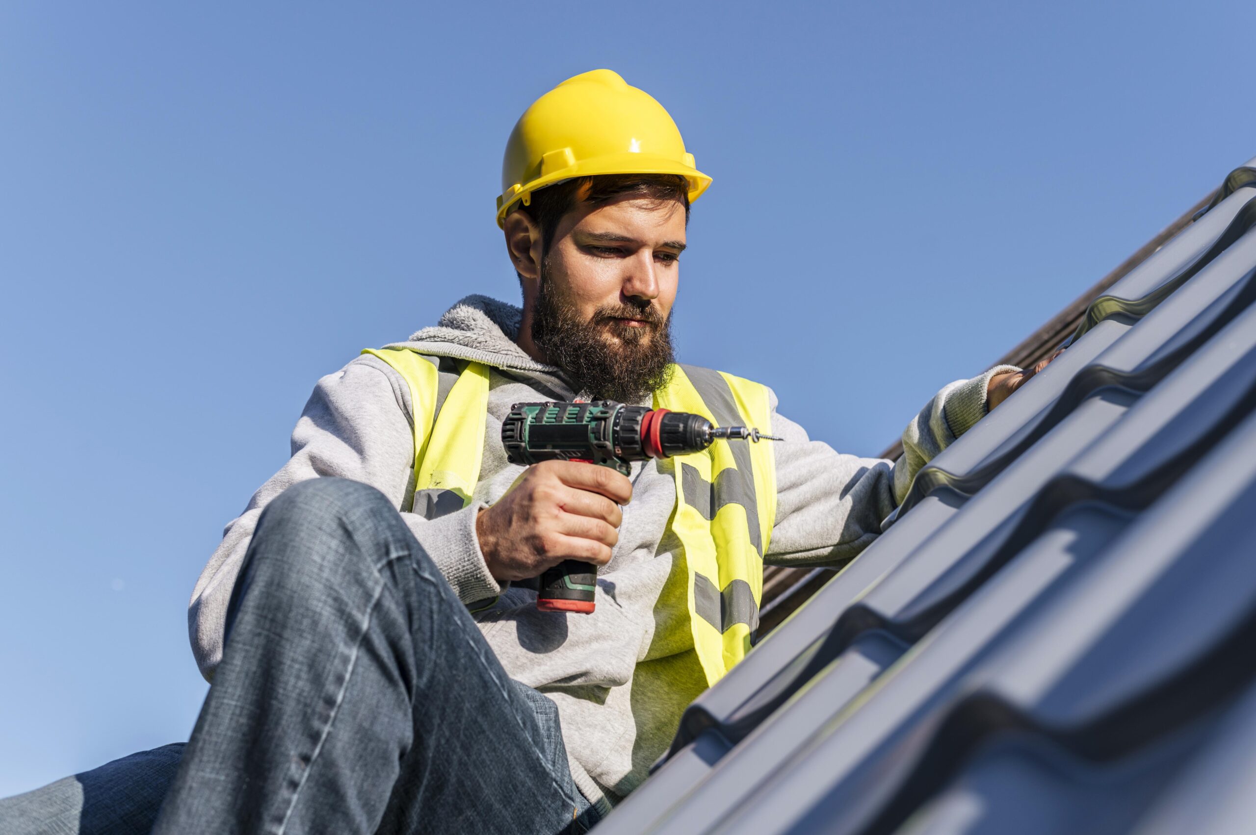 Man in yellow hard hat and safety vest using a power drill on a rooftop, emphasizing professional roof repair services by Tekton Exteriors in Oxford, MS.