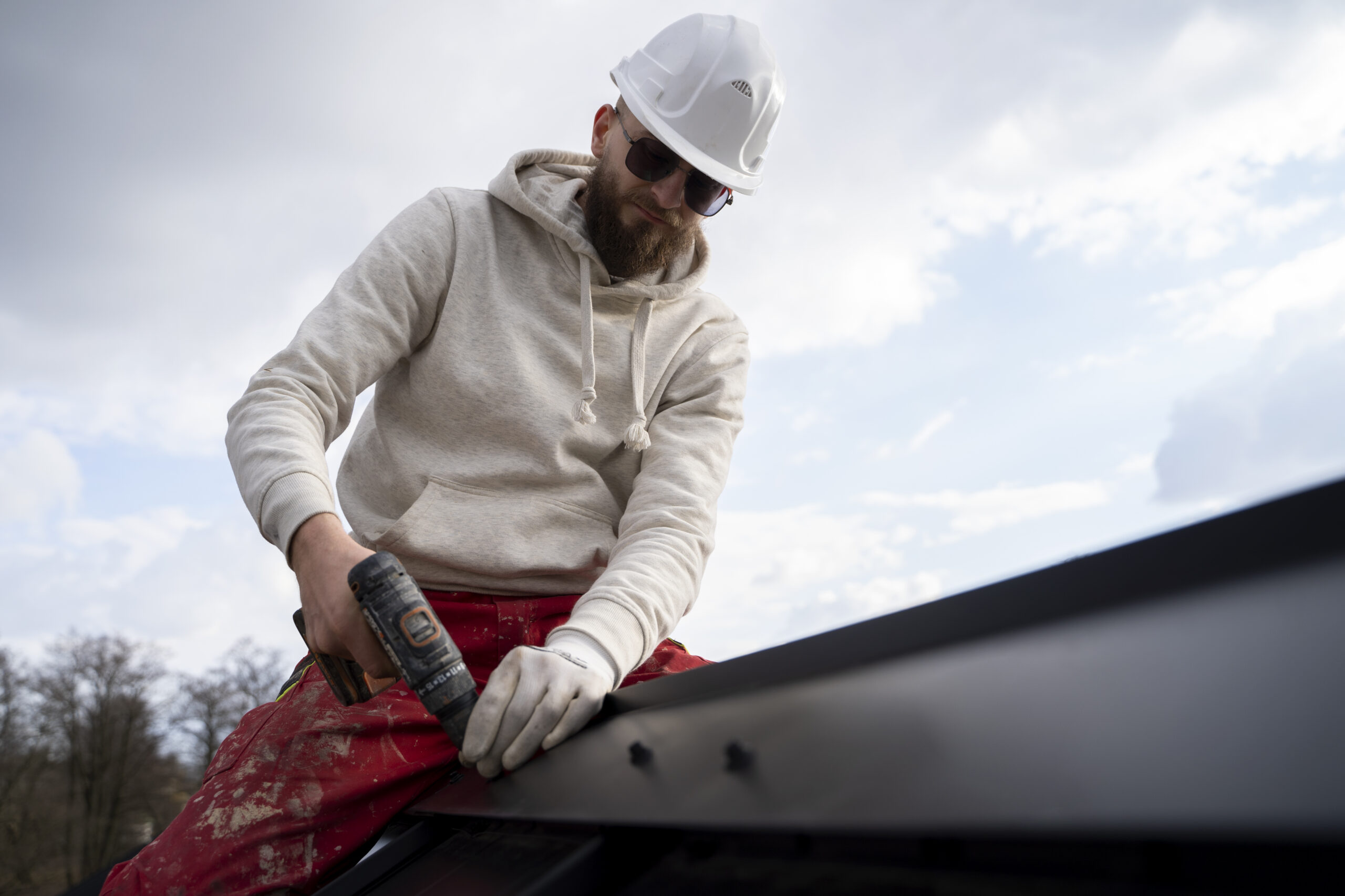 Man wearing a hard hat and sunglasses using a power drill to secure roofing materials, demonstrating roofing repair techniques relevant to emergency roof maintenance after storm damage.