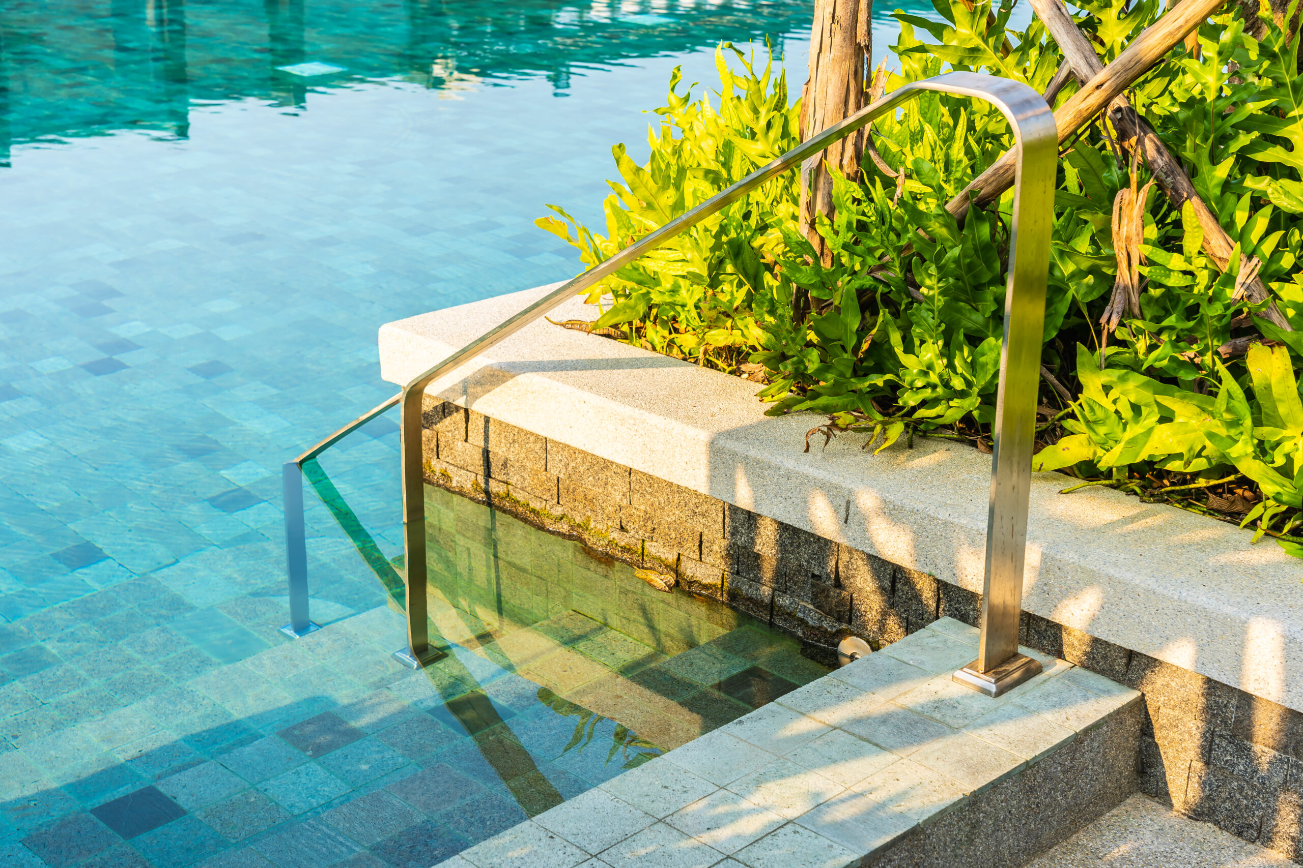 Outdoor swimming pool with a stainless steel stair ladder, surrounded by lush greenery and stone pavers, emphasizing elegant hardscape design.