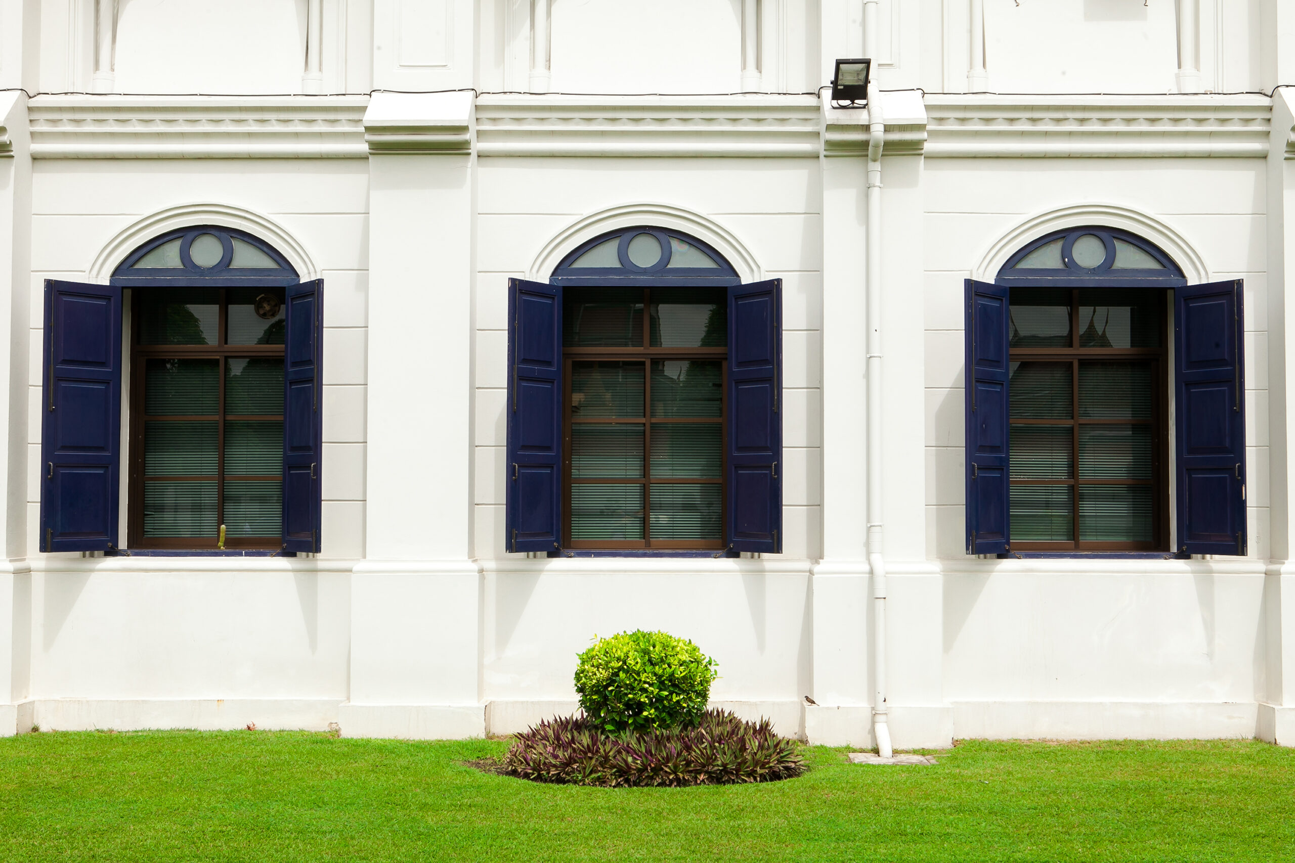 Pella Bow Windows with blue shutters on a white building, surrounded by green grass and decorative plants, illustrating elegant home enhancement by Tekton Exteriors Mississippi.
