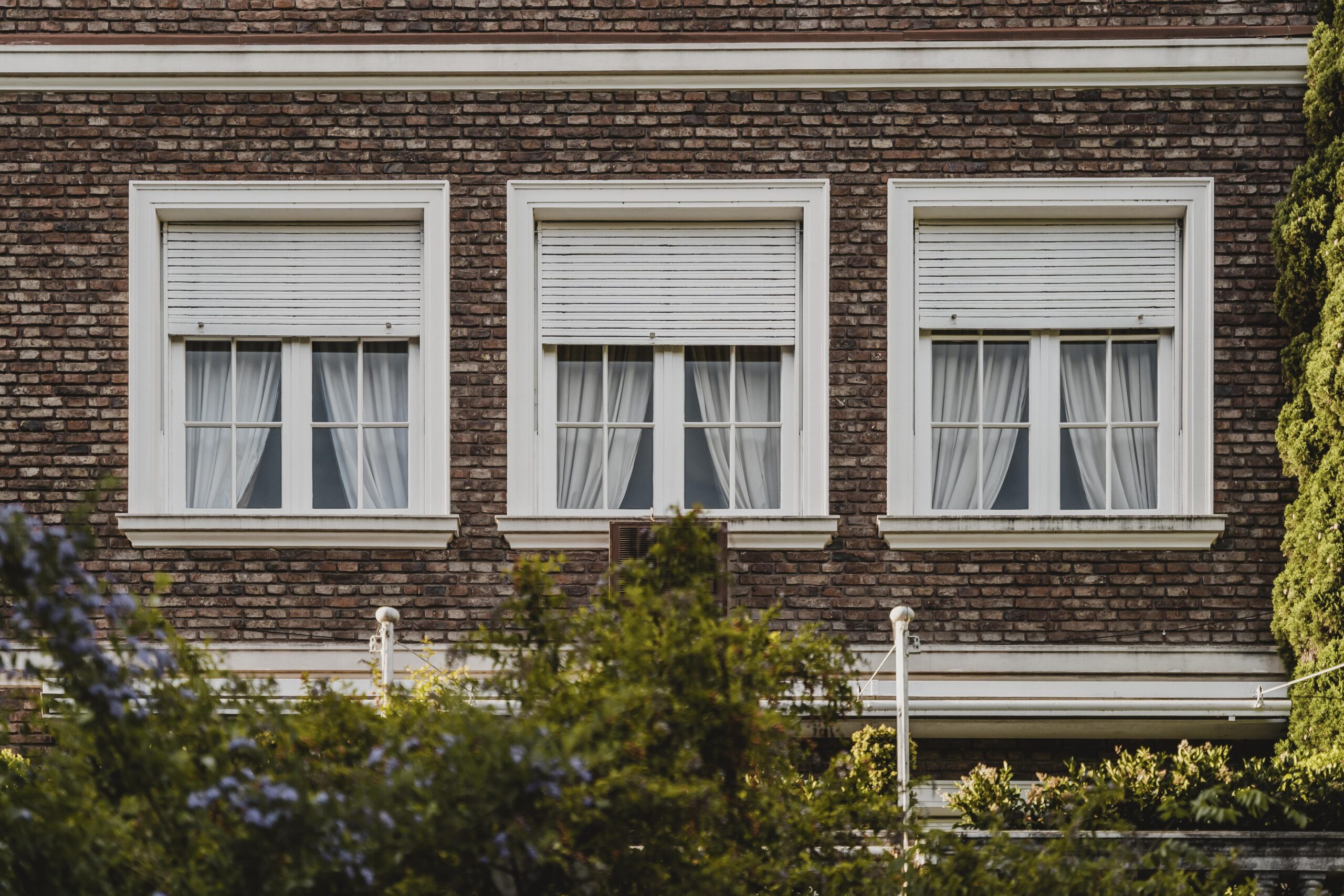 Three Pella Bifold Windows with white frames and sheer curtains, set against a textured brick wall, surrounded by greenery, showcasing a stylish and functional design for enhancing indoor-outdoor spaces.