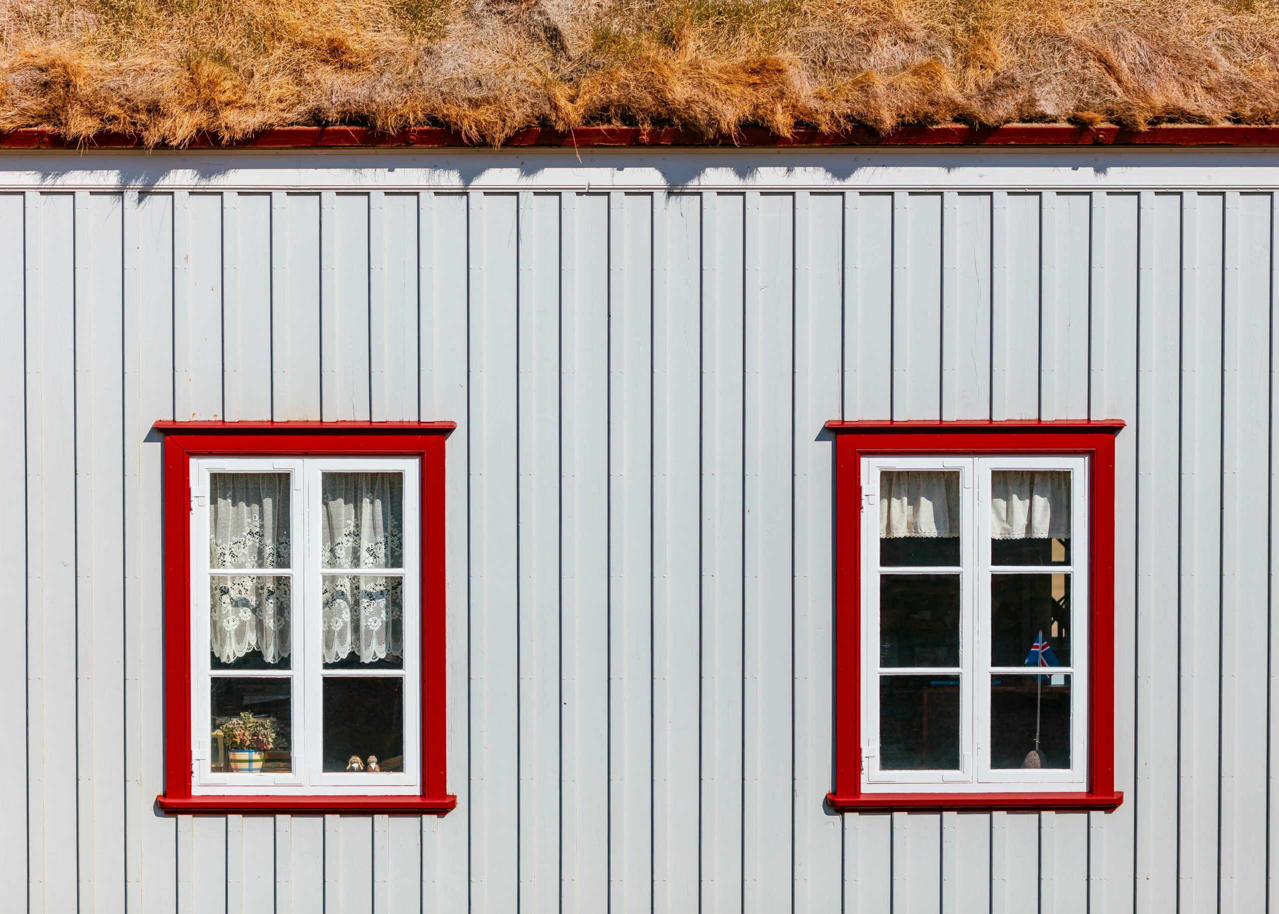 White wooden house exterior featuring two red-framed windows with lace curtains, showcasing a charming and aesthetic design relevant to home improvement and window replacement themes.